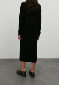Robe noire en tricot côtelé avec des manches longues, une coupe loose et une longueur mi-mollet. Associée à des chaussures noires à semelles épaisses. Design minimaliste.