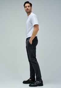 Uomo in camicia bianca a maniche corte, pantaloni neri e mocassini neri in piedi con le mani in tasca, che guarda leggermente di lato su uno sfondo neutro.
