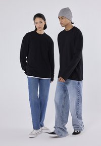 Două modele stau împreună îmbrăcate în tricouri negre cu mânecă lungă, blugi și sneakers. Unul dintre modele poartă o căciulă tricotată gri.