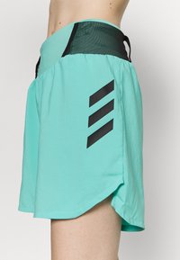 Pantalones cortos deportivos turquesa con cinturilla de malla negra y tres rayas diagonales negras en el lado, llevados en la cintura y el muslo de una persona.