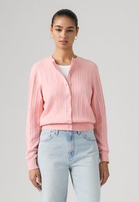 Γυναίκα που φοράει ένα ανοιχτό ροζ πλεκτό ζακέτα με κουμπιά πάνω από λευκή μπλούζα, συνδυασμένο με ανοιχτό μπλε τζιν ψηλής μέσης, στέκεται μπροστά σε γκρι φόντο.