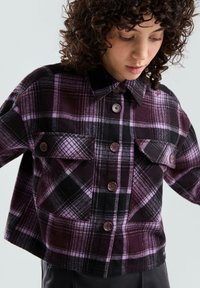 Przycięta koszula w kratę w ciemnym fiolecie i czerni, z guzikami, kołnierzem oraz dwoma kieszeniami na piersi z klapkami. Miękka tekstura tkaniny.