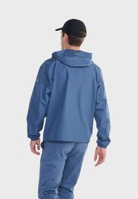 Giacca impermeabile blu con cappuccio, polsini elasticizzati e vestibilità rilassata. Indossata con pantaloni blu coordianti, vista da dietro.