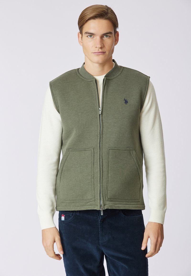 Gilet zipato verde oliva in un tessuto morbido, con due tasche frontali e colletto rialzato, indossato sopra una camicia a maniche lunghe color crema.