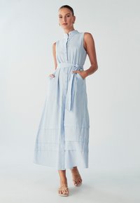 Šviesiai mėlyna be rankovių suknelė su aukštu apykakle, priekiniais sagų, surišamu juosmeniu ir sluoksniuotu kraštu. Turima šoninių kišenių ir lygus audinys.