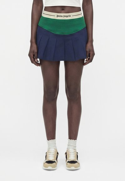 Personne à la peau foncée portant une mini-jupe plissée verte et bleu marine avec une ceinture crème estampillée "Palm Angels", des chaussettes blanches et des baskets beiges.
