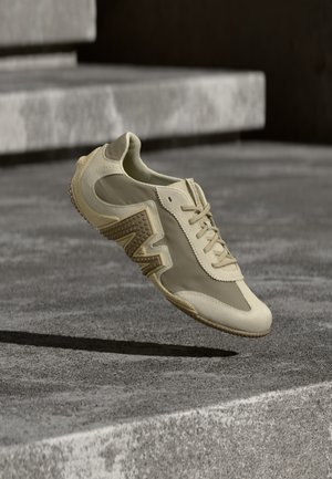 Chaussure de sport beige avec un panneau latéral texturé, des lacets et un bout arrondi, suspendue au-dessus d'une surface en béton avec des ombres contrastées.