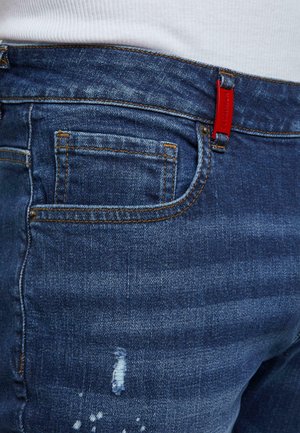 Lähivõte sinistest teksapükstest, millel on oranž õmblus, väike kulumisdetail ning punane brändi silt vööloopi peal.