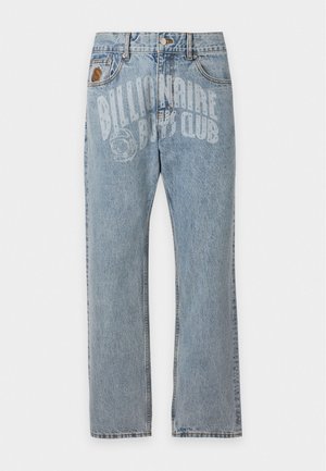 Uomo in piedi che indossa una polo bianca e jeans azzurri chiari con grande testo sbiadito davanti, abbinati a scarpe da ginnastica beige.