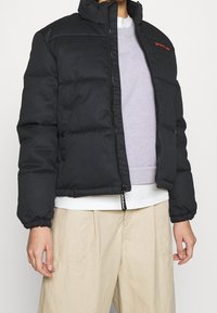 Doudoune noire avec un col montant, fermeture éclair, poignets élastiques, et un petit logo rouge. Portée sur un pull gris et une chemise blanche.