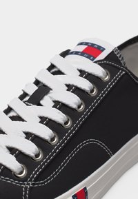 Κοντινό πλάνο μαύρου αθλητικού παπουτσιού με λευκή ραφή και κορδόνια, με το λογότυπο Tommy Jeans σε κόκκινο, λευκό και μπλε στο γλώσσα και την πλευρά.