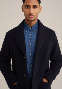Marineblauwe wollen blendjas met een brede kraag, dubbele knoopsluiting aan de voorkant en zijzakken, gedragen over een lichtblauw denim overhemd met knopen.