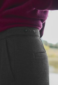 Grijze, gestructureerde broek met een aangesloten taille, voorzien van een zijzak, twee metalen knopen en zichtbare stiksels. Draagt een bordeauxrode trui.