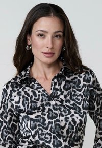 Una donna con lunghi capelli mossi indossa una camicia a stampa leopardata bianca e nera, sfoggiando orecchini a cerchio argentati e un trucco sobrio.