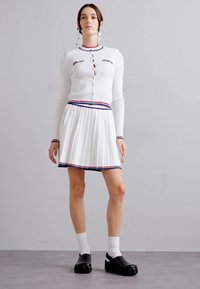 Cardigan blanc en tricot avec col rond, présentant une fermeture à boutons et des accents marine/rouge, associé à une jupe plissée blanche avec une bordure assortie. Sabots noirs.