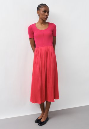 Femeie cu păr împletit stă purtând un top tricotat roz cu mâneci scurte și o fustă midi plisată coral, cu pantofi negri fără toc.