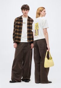 Νεαρός άνδρας και γυναίκα που φοράνε φαρδιά καφέ παντελόνια και λευκά μπλουζάκια με σχέδια· η γυναίκα κρατάει κίτρινη τσάντα, ο άνδρας φοράει καφέ πουκάμισο με καρό.