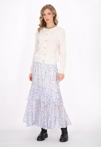 Cardigan crem cu nasturi mari, guler cu volane; fustă în floare, stratificată și fluidă, cu imprimeu floral de liliac; cizme negre la gleznă cu tălpi groase.