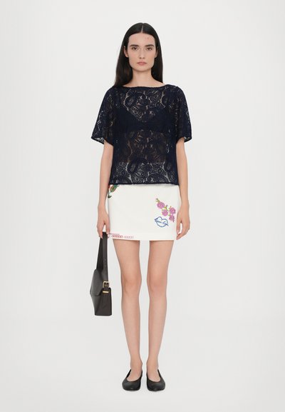 Lace navy top with a loose fit, white mini skirt featuring floral and bird embroidery, black flats, and a structured black bag.