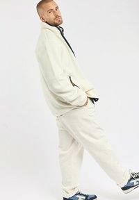 Fleecejacke in Off-White mit hohem Kragen und navyblauen Akzenten, kombiniert mit passenden Off-White-Hosen und blauen Sneakers. Glatte Textur.