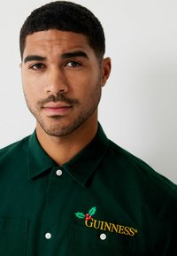 Grüne Baumwollhemd mit einem Button-Down-Kragen, verziert mit gestickten Stechpalmen und dem Wort "GUINNESS" in Gelb auf der linken Seite.