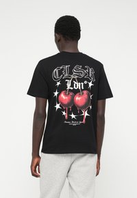 Μαύρο βαμβακερό t-shirt με γραφικό σχέδιο δύο κόκκινων κρεμαστών κερασιών και λευκών αστερίσκων στο πίσω μέρος. Κοντά μανίκια, χαλαρή εφαρμογή.