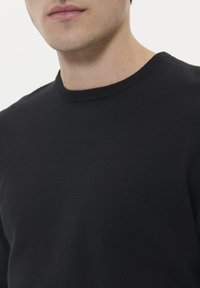 Mies, joka on pukeutunut yksinkertaiseen mustaan pyöreäkaulaiseen neuleeseen, näyttäen alaleukansa ja ylävartalonsa valkoista taustaa vasten.