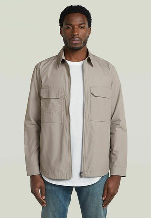 Homme debout portant une veste beige zippée sur une chemise blanche et un jean bleu sur un fond clair uni.