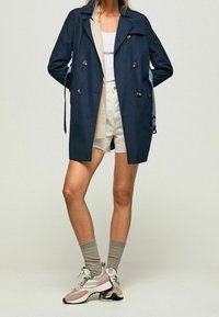 Marinblå trench coat med guldbuttons, med en lös passform och sidofickor. Matchad med en vit linne, beiga shorts och flerfärgade sneakers.