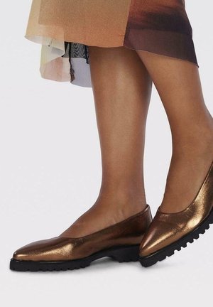 Chaussures plates métalliques marron avec semelles noires texturées portées avec une jupe multicolore, montrant les jambes inférieures sur fond blanc.