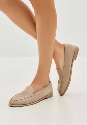 Mocassini beige senza lacci con una morbida texture in suede, punta arrotondata, cuciture ridotte al minimo e una suola in gomma bassa, mostrati su gambe nude.