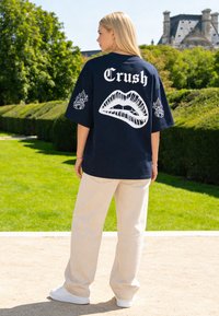 Donna in piedi all'aperto che indossa una t-shirt oversize blu navy con la scritta "Crush" e una grafica di labbra sul retro, pantaloni beige e scarpe da ginnastica bianche.