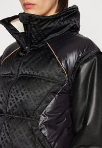 Schwarze Daunenjacke mit strukturiertem Muster, glänzenden schwarzen Details, hohem Kragen und schwarzen Lederärmeln mit Kontrastnähten.
