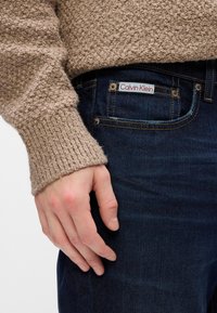 Beige textúrájú pulóver bordás mandzsettákkal, sötétkék farmernadrágon viselve, amelyen a hátsó zsebnél egy piros és fehér "Calvin Klein" címke található.