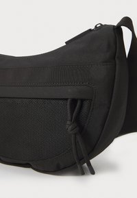 Schwarze Umhängetasche aus strapazierfähigem Stoff, mit einer Netz-Vordertasche, verstellbarem Tragegurt und einem Reißverschluss. Glatte Textur überall.