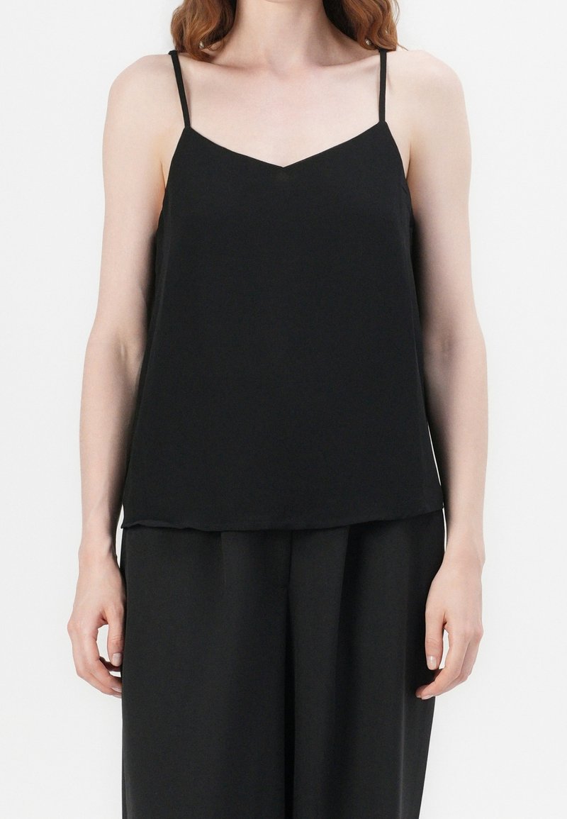 Femme portant un haut noir sans manches à fines bretelles et un pantalon noir ample, debout devant un fond blanc.