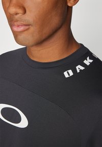 Svart treningstrøye med teksturert stoff, rund hals og hvite logoaksenter på brystet og skuldrene.