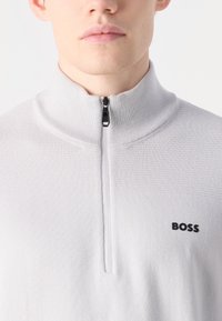 Lys grå zip-up genser med høy krage, teksturert stoff, og svart "BOSS"-logo øverst til venstre på brystet. Glidelåsen er svart med metalltrekk.