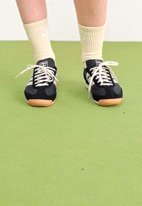 Juodi sportiniai batai su baltais akcentais ir guminiu padu, derinami su šviesiais smėlio spalvos kompresiniais kojiniais. Paviršius yra lygus žalias tekstūras.