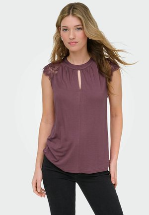 Młoda kobieta o długich blond włosach, ubrana w fioletowy top bez rękawów z koronkowymi ramionami i czarne spodnie na jednolitym tle.