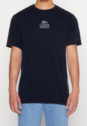 Mann trägt ein schwarzes Lacoste-T-Shirt mit grünem Krokodil-Logo und Schriftzug, kombiniert mit hellblauen Jeans.