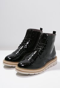 Botas de tobillo de charol negro con detalles de brogue, parte delantera con cordones y suela de goma blanca. Presentan una textura suave y un diseño estructurado.