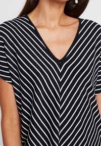 Camiseta de rayas blancas y negras con cuello en V, mangas cortas y un patrón en forma de chevrón, confeccionada en una tela ligera y texturizada.