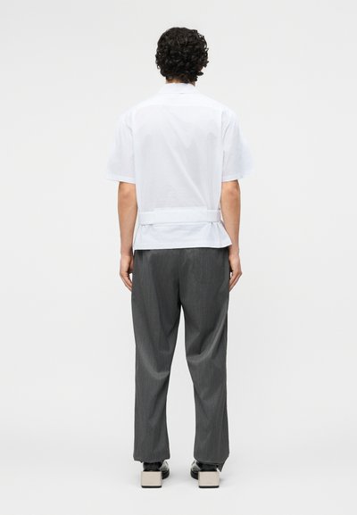 Άνδρας με σγουρά μαλλιά που φοράει λευκό πουκάμισο με κοντά μανίκια και γκρι παντελόνι, στέκεται με την πλάτη προς το μέρος, μπροστά σε ένα απλό φόντο.