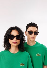 En kvinde iført sorte cat-eye solbriller med en blank finish, og en mand med dybe ambersfarvede rektangulære solbriller med matte detaljer. Begge bærer grønne t-shirts.