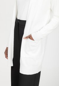 Witte cardigan met open voorkant, lange mouwen en een getextureerde afwerking. Beschikt over een zak aan de zijkant. Gecombineerd met zwarte broek.
