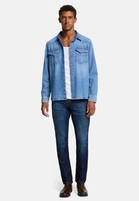Lichtblauwe denim overhemd met twee borstzakken, knoopsluiting aan de voorkant en spitse kraag, gecombineerd met donkerblauwe jeans en bruine enkellaarsjes.