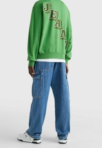 Zelená mikina s kontrastním nápisem "JEAN" na zádech, doplněná volnými modrými džínovými cargo kalhotami a bílými teniskami.