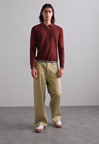 Un giovane uomo indossa un polo a maniche lunghe bordeaux, pantaloni larghi beige e sneakers bianche su un pavimento grigio di fronte a una parete semplice.
