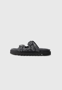Black sandal with three adjustable straps, white stitching, decorative metal details, and thick jagged sole on a white background.